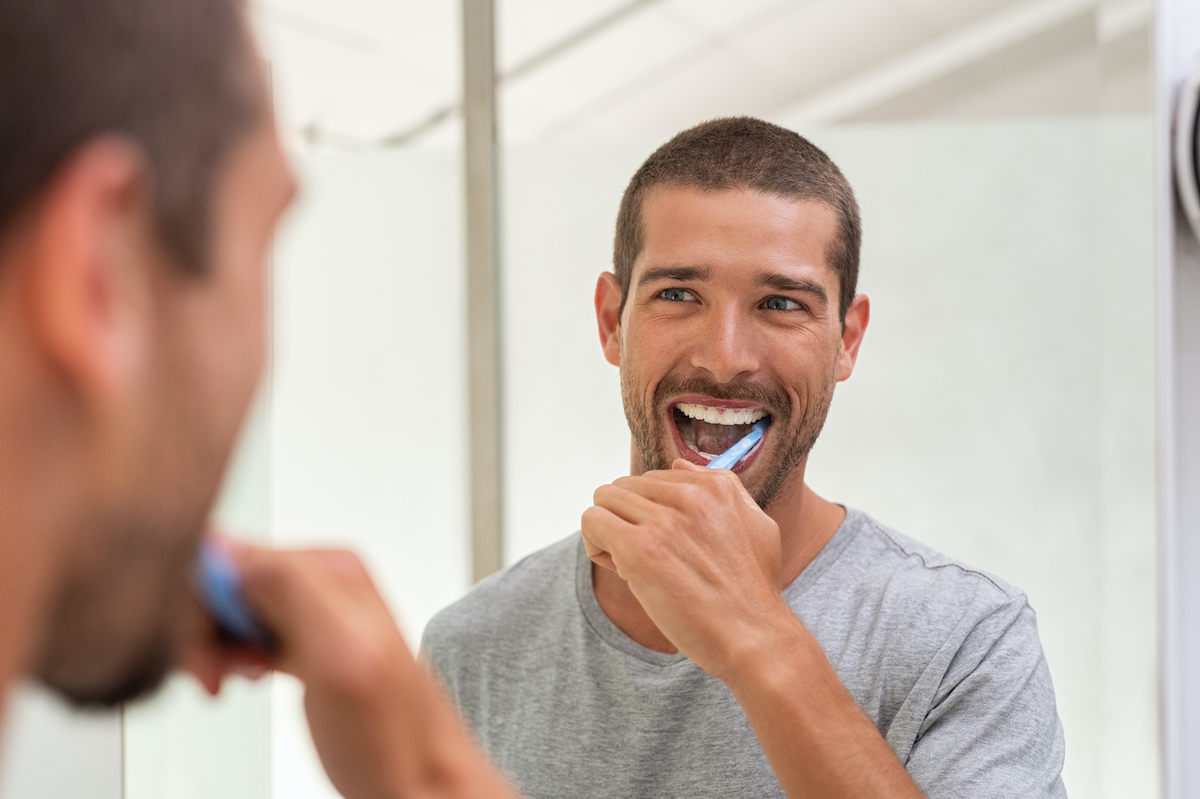 Smiling young man brushes his teeth while looking at himself in the bathroom mirror